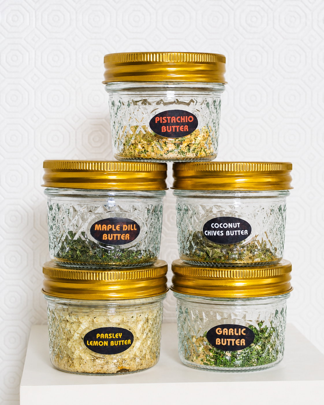 Five jars of different types of butter with gold lids on a white background